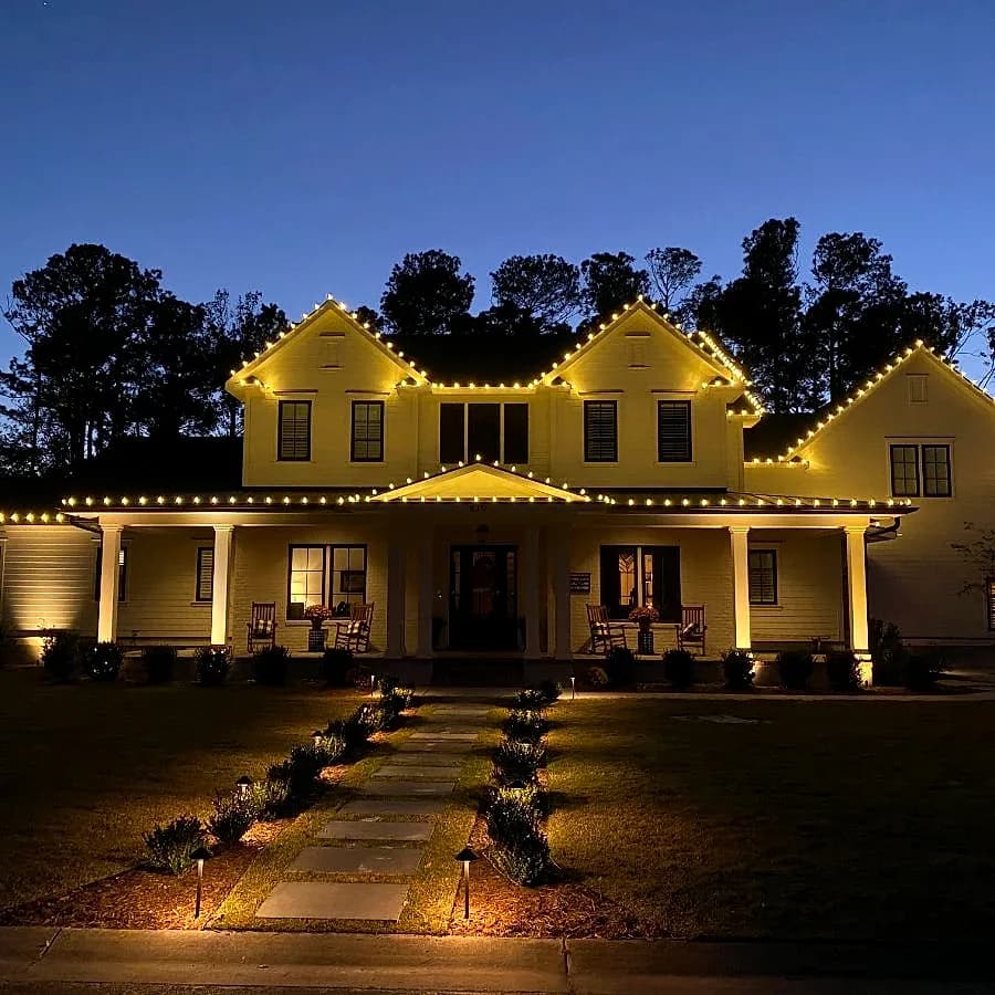 Warm white on a white farmhouse — permanent outdoor lighting installed by Buzz Lighting's crew